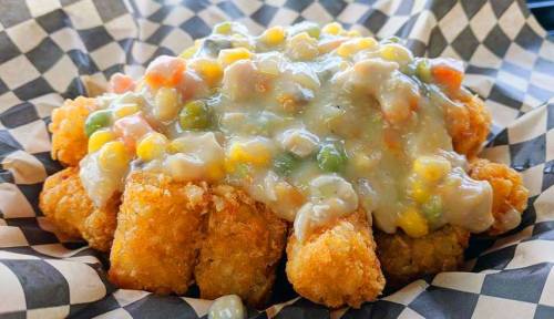 A plate of crispy tater tots topped with creamy vegetable gravy and mixed vegetables, served in a checkered paper basket.