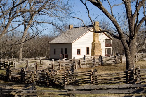 A historic white house with a stone chimney, surrounded by a wooden fence and bare trees in a rural setting.