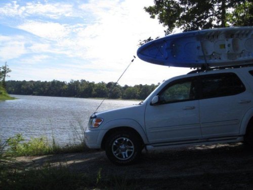 A white SUV parked by a calm lake, with a blue kayak secured on its roof under a clear sky.