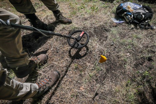 A person using a metal detector on the ground, with a yellow flag marking a spot nearby.