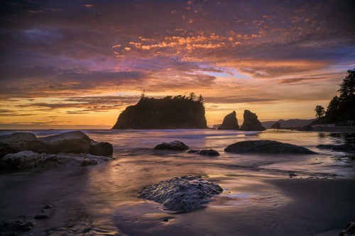 Sunset over a rocky beach with silhouettes of cliffs and trees, reflecting colors in the calm water.