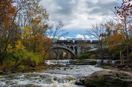 A train crosses a stone bridge over a river, surrounded by autumn trees and a cloudy sky. Water cascades below.