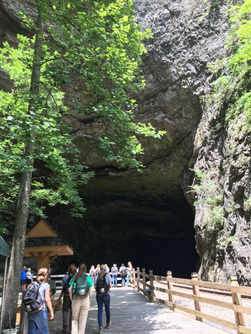 Visitors walk along a wooden path leading to a large cave entrance, surrounded by lush greenery and rocky cliffs.