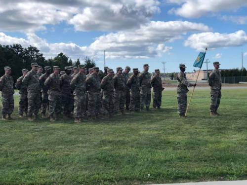 A group of soldiers in military uniforms standing in formation, with one holding a flag, under a cloudy sky.
