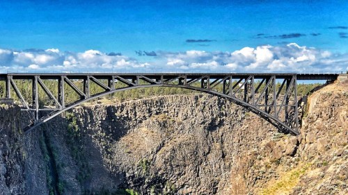 A large black metal arch bridge spans a deep canyon under a blue sky with scattered clouds.