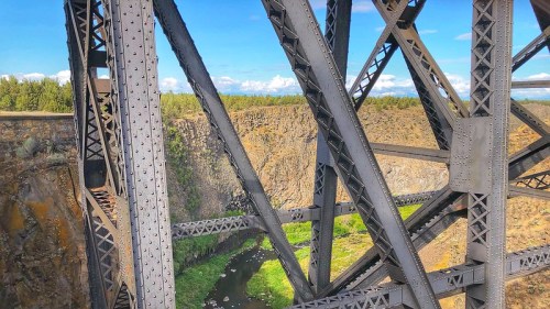 View of a metal bridge structure over a canyon with a river below and trees in the background under a blue sky.