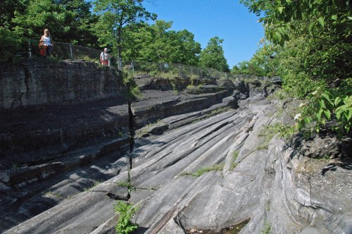 Two people stand on a rocky ledge overlooking a deep, eroded ravine surrounded by greenery under a clear blue sky.
