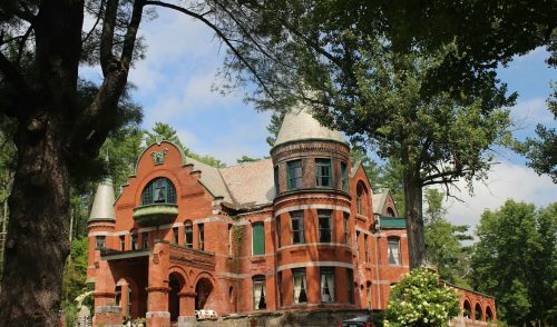 A large, ornate red brick mansion with turrets, surrounded by tall trees and greenery under a blue sky.