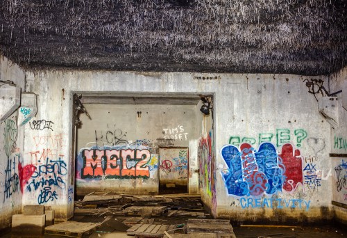 Graffiti-covered walls in a dimly lit, abandoned room with debris on the floor and a textured ceiling.