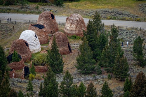 Historic beehive kilns surrounded by trees, with a road and two people walking nearby.