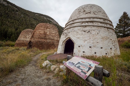Historic brick kilns with a sign in front, surrounded by grass and mountains under a cloudy sky.