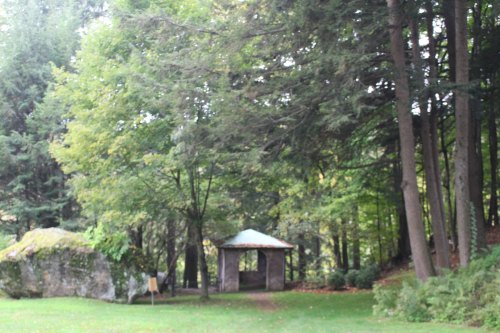 A serene forest scene featuring tall trees, a small gazebo, and a large rock in a grassy area.