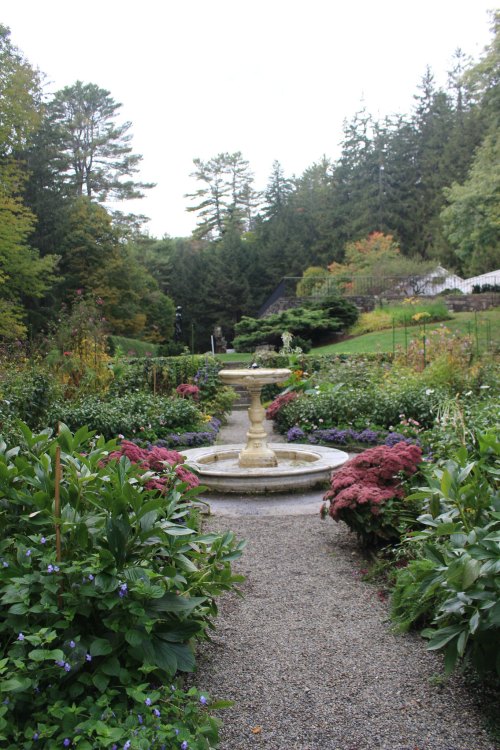 A serene garden path lined with colorful flowers leads to a central fountain, surrounded by lush greenery and trees.