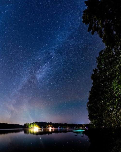 A serene night sky filled with stars and the Milky Way, reflecting on a calm lake surrounded by trees.
