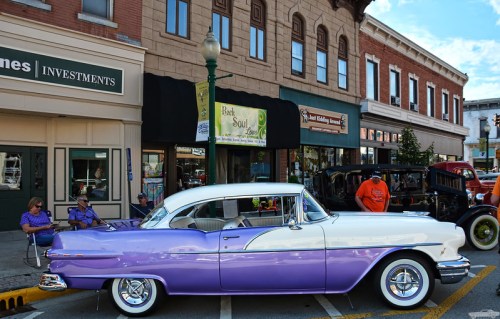 A vintage purple and white car parked on a street with people sitting nearby and historic buildings in the background.
