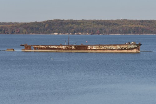 A rusted, abandoned barge partially submerged in calm water, with trees in the background and a clear blue sky.