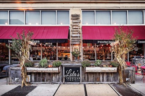 Exterior of Lela's European Café with wooden planters, fall decor, and red awnings, showcasing a cozy atmosphere.