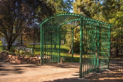 A green metal gate opens to a lush garden with trees and flowers, welcoming visitors to a serene outdoor space.