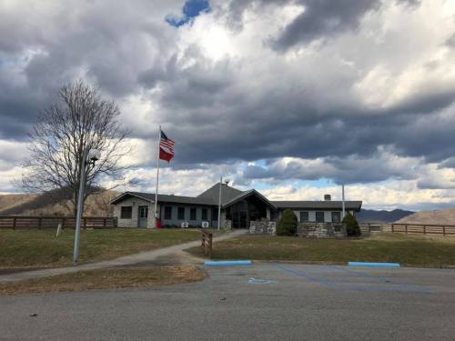 A stone building with a flag in front, set against a cloudy sky and rolling hills in the background.