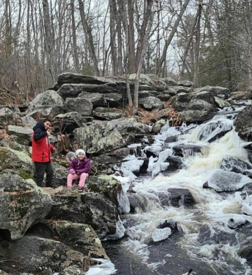 Two children by a rocky stream, one standing with a stick and the other sitting on a rock, surrounded by trees.