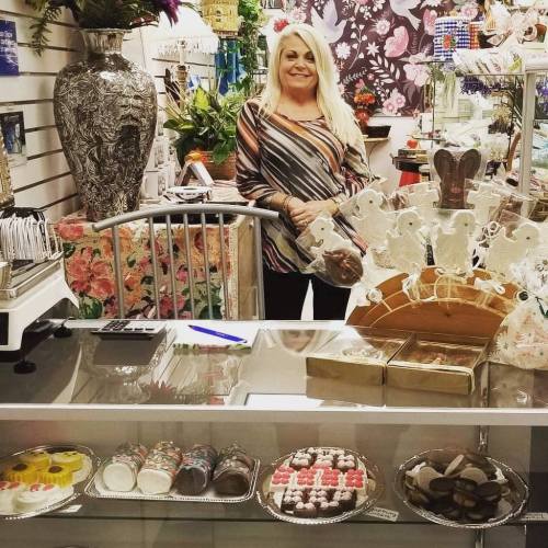 A smiling woman stands behind a display of assorted baked goods in a colorful shop filled with decorations.