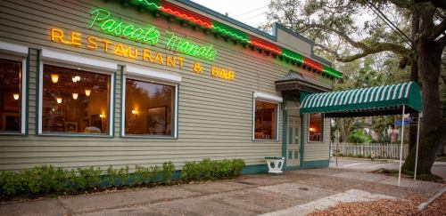 Exterior of Pascal's Manale restaurant, featuring neon signage and a green-striped awning, surrounded by trees.