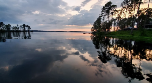 A serene lake at sunset, with trees reflecting on the calm water and a cloudy sky above.