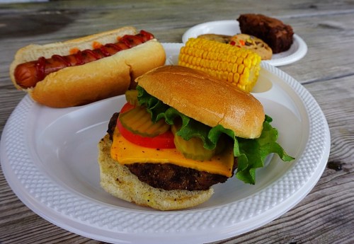 A plate with a cheeseburger, hot dog, corn on the cob, and two cookies, all on a wooden table.