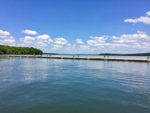 A serene lake scene with calm water, a blue sky, fluffy clouds, and a wooden dock extending into the water.