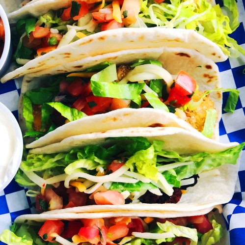 Three tacos filled with lettuce, tomatoes, cheese, and grilled ingredients, served on a blue checkered tray.