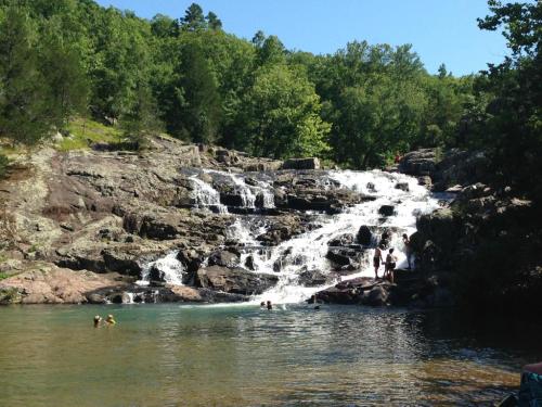A scenic waterfall cascades over rocks into a clear pool, with people swimming and enjoying the sunny day.