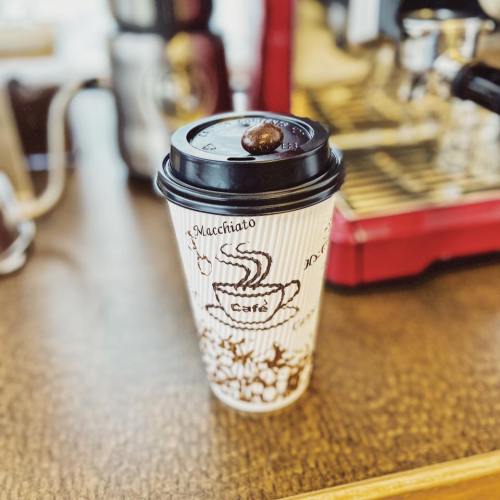 A coffee cup with a macchiato design, topped with a coffee bean, sits on a wooden table near an espresso machine.
