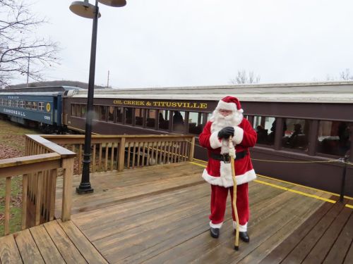Santa Claus in a red suit stands on a wooden platform near a vintage train car labeled "Oil Creek & Titusville."