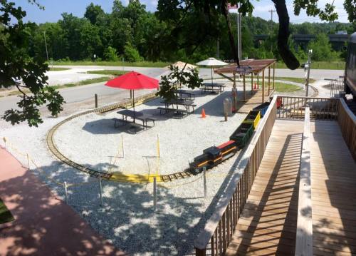 A miniature train track with a small train, picnic tables, and umbrellas in a gravel area surrounded by trees.