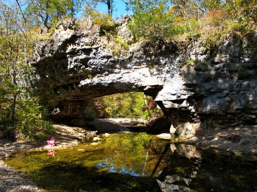 A natural stone arch over a clear stream, surrounded by trees and colorful autumn foliage.