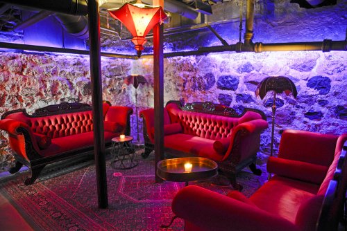 Cozy underground lounge with red velvet sofas, a decorative lamp, and stone walls illuminated by purple lighting.