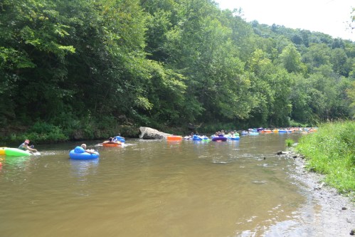 People floating on colorful inner tubes down a calm river, surrounded by lush green trees and rocky banks.