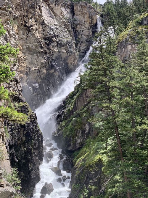 A cascading waterfall flows down rocky cliffs, surrounded by lush green trees and rugged terrain.
