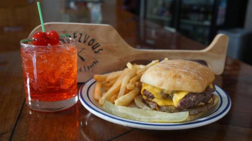 A cheeseburger with fries and a cherry soda on a wooden table, with a paddle-shaped sign in the background.
