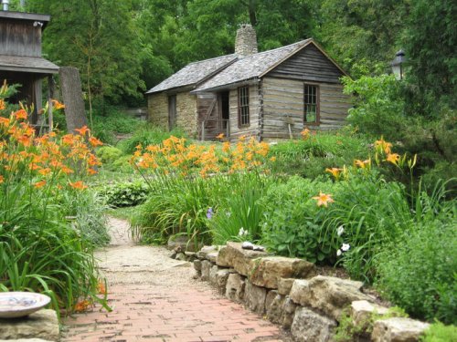 A rustic wooden cabin surrounded by vibrant orange flowers and lush greenery, with a stone pathway leading to it.