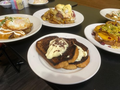 A table with four plates of food, including French toast with cream, eggs, and various potato dishes.