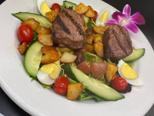A plate of grilled steak on a bed of greens, with potatoes, cucumbers, cherry tomatoes, and garnished with an orchid.
