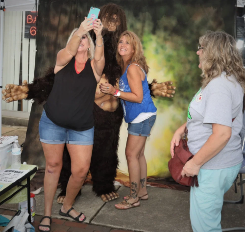 Three women pose for a selfie with a Bigfoot costume in front of a nature-themed backdrop.