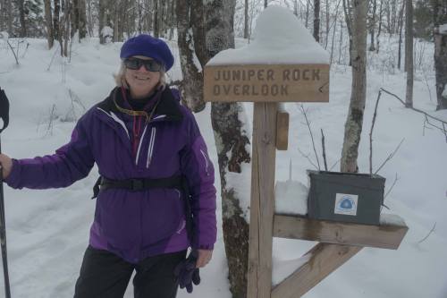 A person in a purple jacket stands next to a sign for Juniper Rock Overlook in a snowy forest.