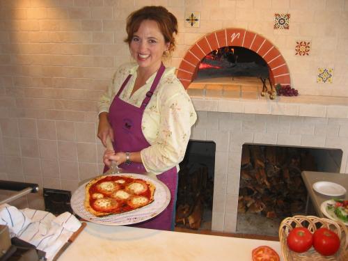 A woman in an apron smiles while holding a freshly baked pizza in a rustic kitchen with a wood-fired oven.
