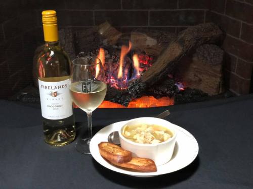 A bottle of white wine and a glass beside a bowl of soup and toasted bread, with a cozy fireplace in the background.