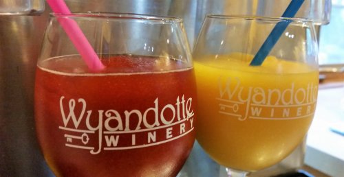 Two glasses of colorful drinks with straws, featuring the Wyandotte Winery logo. One is red and the other is yellow.