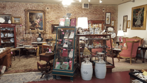 A vintage shop interior featuring antique furniture, decorative items, and a display of collectibles against a brick wall.