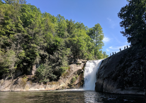 A waterfall cascades down rocky cliffs surrounded by lush green trees, with silhouettes of people at the top.