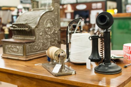 A vintage display featuring an antique cash register, telephone, spool of thread, and other nostalgic items on a wooden table.